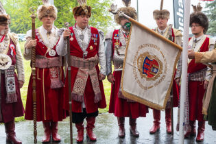 Siedmiu członków Bractwa Kurkowego w tradycyjnych strojach historycznych. Jeden trzyma proporzec z napisem „Societis Jaculatorum Cracoviensis – Semper Fidelis”, a drugi srebrnego kura – symbol Bractwa