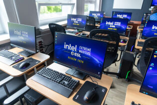 Nowa sala komputerowa w szkole. Niebieskie pomieszczenie z kilkunastoma stanowiskami komputerowymi. Jasne stoliki. na ekranach napis Intel Extreme Master.    