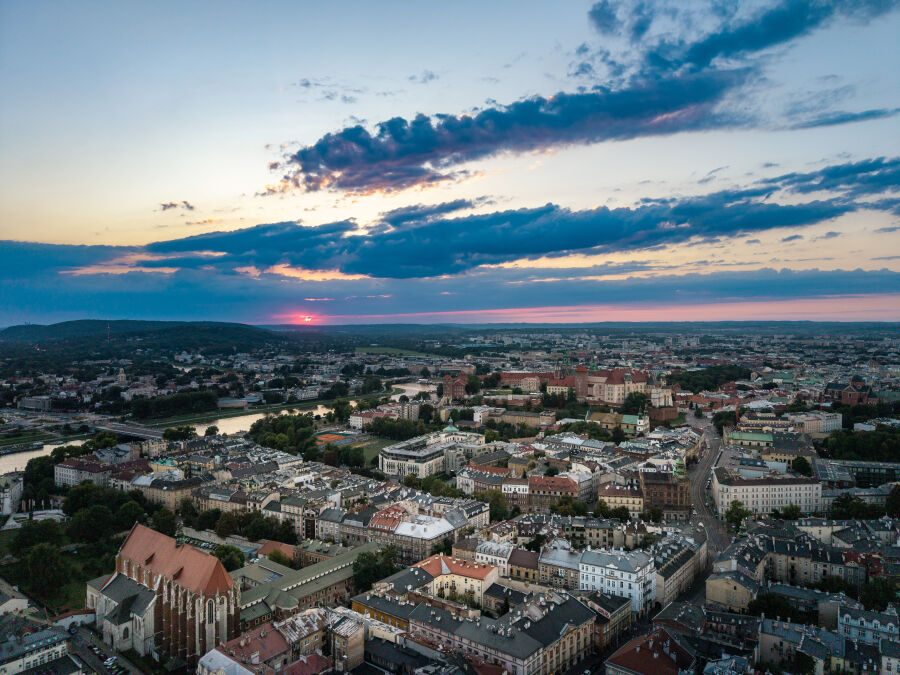 Zdjęcie Krakowa wykonane z drona. Widoczne jest niebo z zachodzącym słońcem oraz zwarta zabudowa miejska centrum i przestrzenie zieleni na jego obżerzach.  