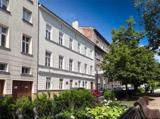 Budynek mieszkalny z jasną elewacją, otoczony zielenią i drzewami.