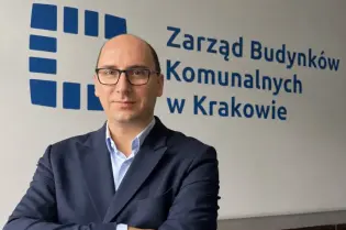 Mężczyzna w garniturze stoi z założonymi rękami przed logo Zarządu Budynków Komunalnych w Krakowie.