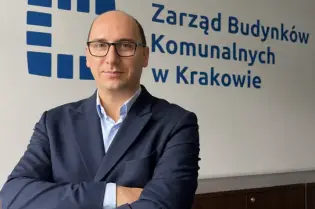 Mężczyzna w garniturze stoi z założonymi rękami przed logo Zarządu Budynków Komunalnych w Krakowie.