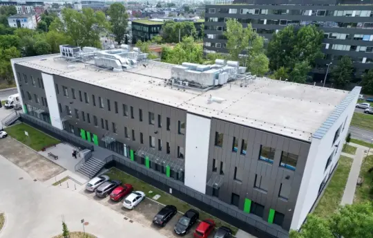 Nowoczesny budynek biurowy z zielonymi detalami i parkingiem.