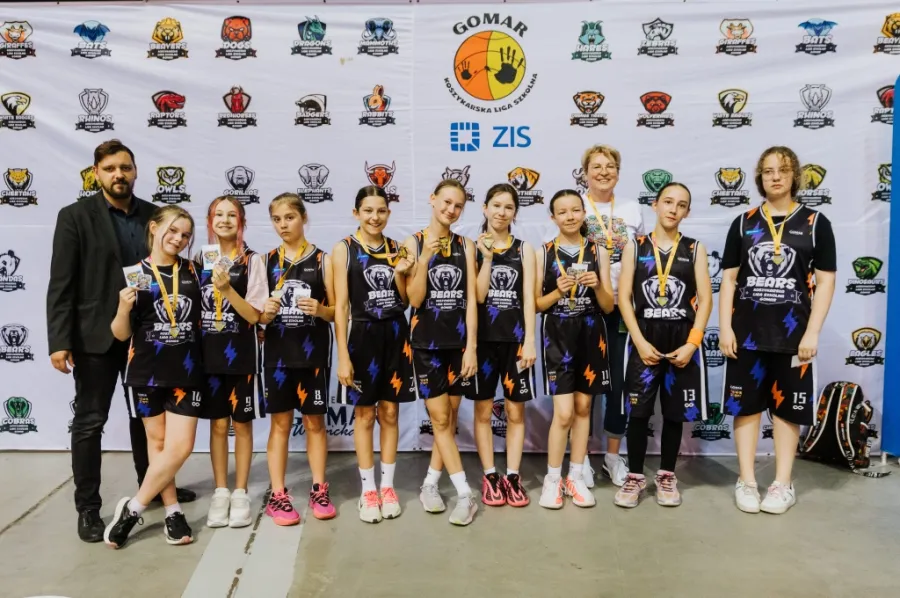 Zespół dziewcząt w strojach koszykarskich z medalami, stojący przed tłem z logo i herbami.