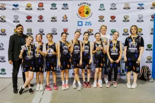 Zespół dziewcząt w strojach koszykarskich z medalami, stojący przed tłem z logo i herbami.