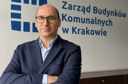 Mężczyzna w garniturze stoi z założonymi rękami przed napisem Zarząd Budynków Komunalnych w Krakowie.