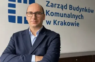 Mężczyzna w garniturze stoi z założonymi rękami przed napisem Zarząd Budynków Komunalnych w Krakowie.