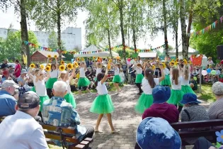 Dzieci tańczą w zielonych spódnicach, trzymając słoneczniki, w parku pełnym widzów.