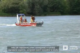 Na zalewie w słoneczny dzień płynie motorówka z ratownikami i przedstawicielami służb. Wokół widoczna jest spokojna tafla wody, trzcinowiska oraz zielona trawa na brzegach zbiornika.