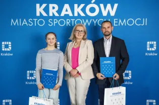 Kobieta i dwoje ludzi trzymających torby z logo Krakowa na tle niebieskiego banera.