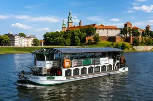 Statek wycieczkowy płynący po rzece z widokiem na zamek i zielone wzgórza.