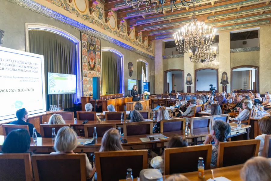 Konferencja w sali z wykładowcą i uczestnikami siedzącymi w rzędach.
