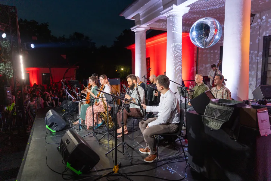 Koncert na świeżym powietrzu z zespołem grającym na scenie, w tle tłum i dekoracyjne oświetlenie.