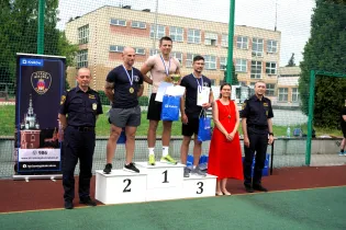 Mężczyźni na podium z medalami, obok kobieta w czerwonej sukience i przedstawiciele służb mundurowych.
