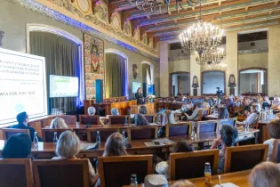 Konferencja z prelegentem, uczestnicy siedzą w sali z dekoracjami i ekranami.