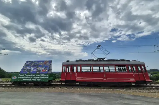 Czerwony tramwaj na torach z plakatem reklamowym w tle.