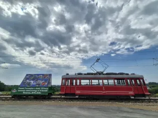 Czerwony tramwaj z reklamą na boku, stojący na torach pod chmurami.
