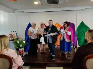 Cztery osoby na scenie, jedna z nich trzyma kartkę, obok kolorowe tkaniny i dekoracje.