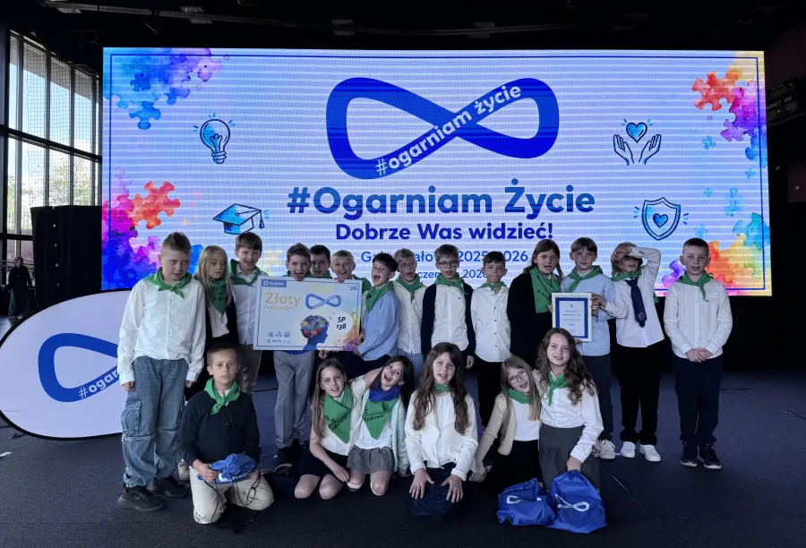 Grupa dzieci w białych koszulach i zielonych chustach, stojąca przed dużym ekranem z napisem #Ogarn iam Życie.