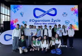 Grupa dzieci w białych koszulach i zielonych chustach, stojąca przed dużym ekranem z napisem #Ogarn iam Życie.