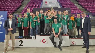Grupa dzieci z medalami i pucharem na podium po zawodach sportowych.