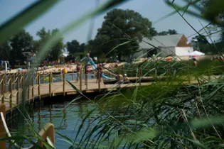 Plaża z pomostem, ludzie wypoczywający nad wodą, otoczenie zieleni.