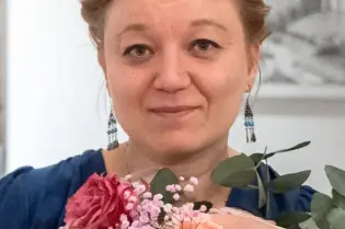 Kobieta trzyma bukiet różowych i białych kwiatów, ma na sobie niebieską bluzkę i kolczyki.