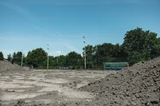 Widać rozkopane boisko, ławki dla zawodników, ogrodzenie a za nimi drzewa.
