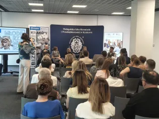 Na pierwszym planie widać uczestników spotkania siedzących tyłem do obiektywu. W głębi sali znajdują się prelegenci, a za nimi ustawiono banery.
