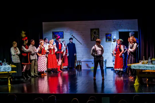 Scena teatralna z aktorami w tradycyjnych strojach, interakcje między postaciami w wiejskim otoczeniu.