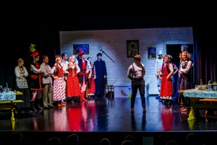 Scena teatralna z aktorami w tradycyjnych strojach, interakcje między postaciami w wiejskim otoczeniu.