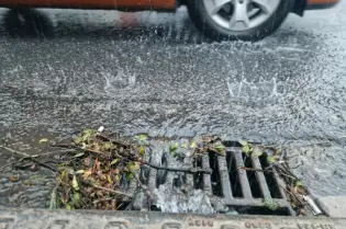 Deszcz spada na ulicę, woda zbiera się wokół kratki ściekowej, obok przejeżdża samochód.