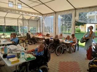 Osoby na wózkach inwalidzkich uczestniczą w zajęciach w namiocie, jedna osoba gra na gitarze.