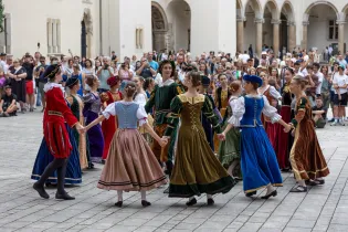 Grupa tancerzy w tradycyjnych strojach ludowych tańczy w kręgu na placu wśród widowni.