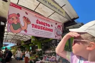 Dziecko w okularach przeciwsłonecznych na tle straganów i baneru zapraszającego na Święto Truskawki.
