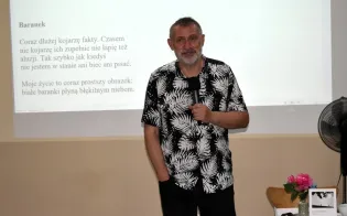 Mężczyzna w koszuli w kwiaty stoi przy mównicy, obok prezentacja na ekranie.