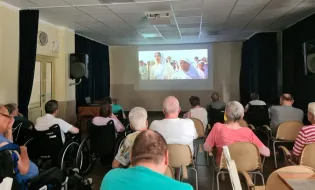 Grupa osób w wózkach inwalidzkich ogląda film na dużym ekranie w sali.