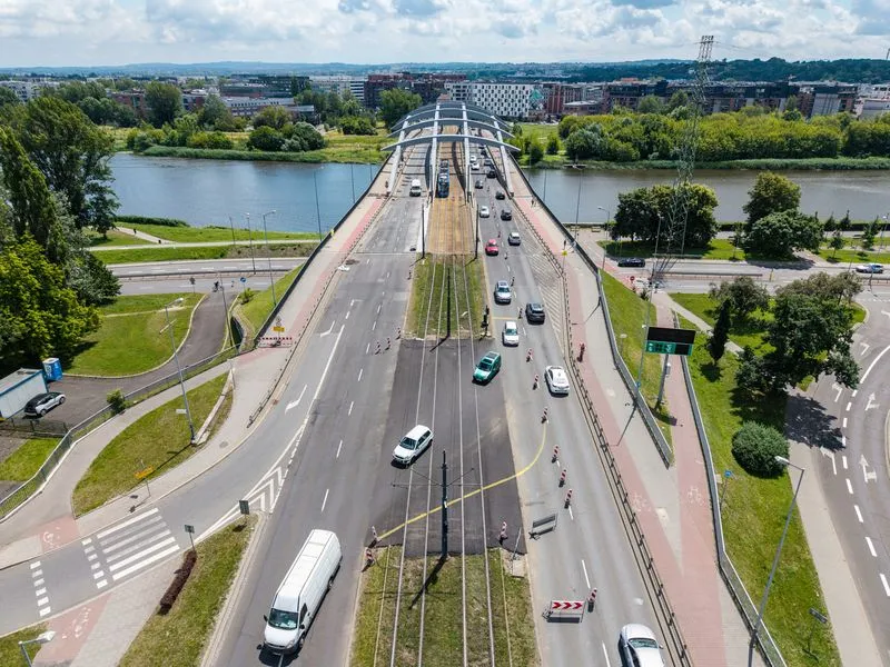 Most nad rzeką z ruchem drogowym i tramwajowym, otoczenie zieleni i zabudowy miejskiej.