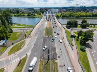 Most nad rzeką z ruchem drogowym i tramwajowym, otoczenie zieleni i zabudowy miejskiej.