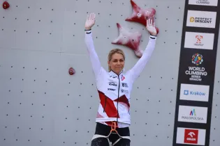 Zawodniczka wspinaczki sportowej z uniesionymi rękami na tle ścianki wspinaczkowej.