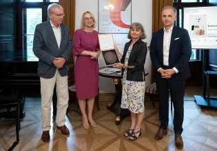 Fotografia wykonana podczas uroczystości wręczenia nagrody im. Karla Dedeciusa w Willi Decjusza 1 lipca 2026 r. Na zdjęciu stoją cztery osoby. Kobieta w środku trzyma otwarte pudełko z medalem.