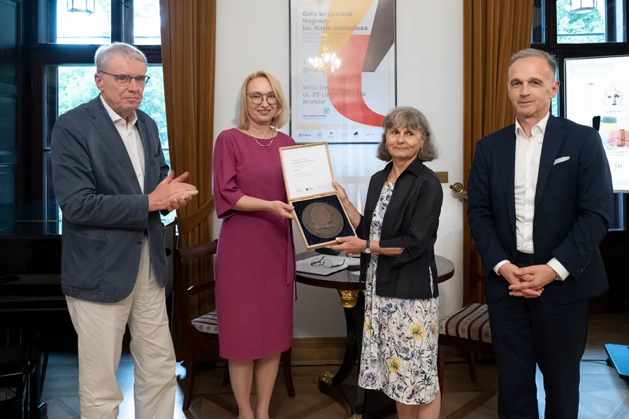 Wręczenie nagrody w eleganckim wnętrzu, cztery osoby w formalnych strojach, jedna trzyma medal i dyplom.
