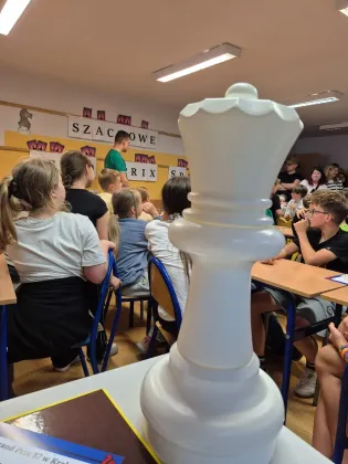 Biały król szachowy w sali lekcyjnej, uczniowie siedzą w tle.