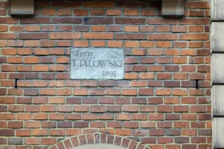 Tablica z napisem Fecit T. Talowski 1891 na tle ceglanej ściany.