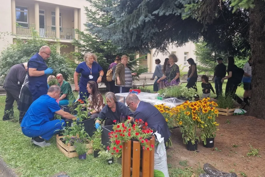 Grupa ludzi sadzi kwiaty w ogrodzie, otoczona zielenią i drewnianymi skrzynkami.
