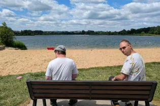 Dwóch mężczyzn siedzących na ławce nad jeziorem, w tle plaża i chmury na niebie.