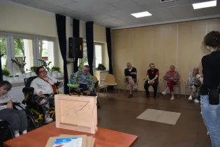 Grupa osób starszych w sali, część z nich siedzi na krzesłach, inne w wózkach inwalidzkich.