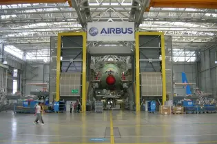 W ogromnej hali przemysłowej na środku widoczny kadłub samolotu, obok niego skomplikowane maszyny oraz krzątający się pracownicy. Nad kadłubem widoczny napis Airbus.  