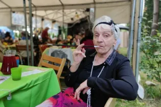 Starsza kobieta w eleganckim stroju, paląca papierosa, siedzi przy stole w ogrodzie.