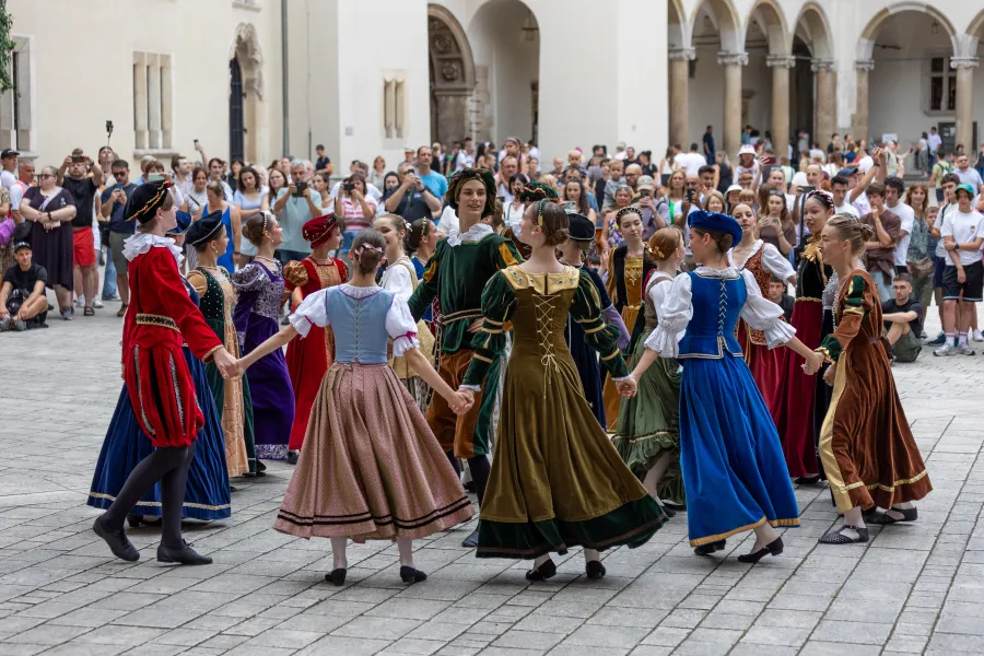 Grupa tancerzy w tradycyjnych strojach ludowych tańczy w kręgu na placu wśród widowni.